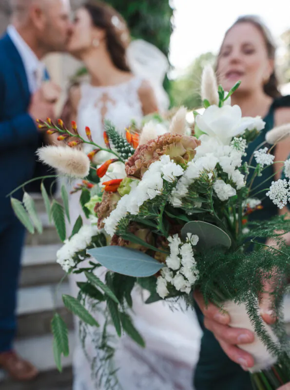 A close-up shot of a textured wedding bouquet featuring white flowers, greenery, and bunny tails, with a bride and groom kissing softly in the blurred background.