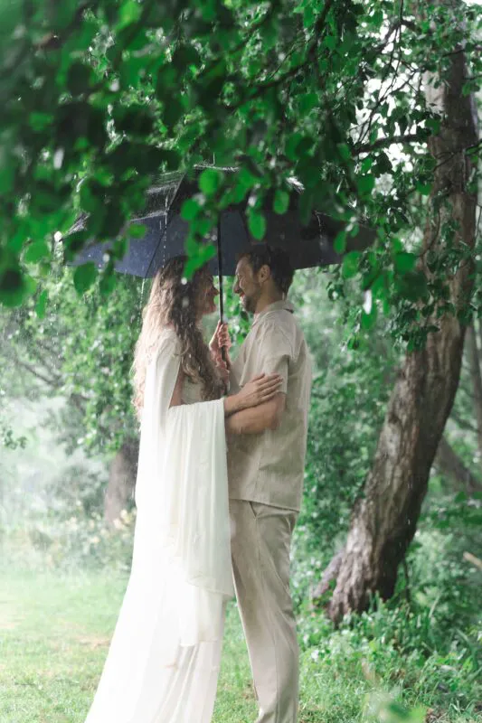 A newlywed couple shares an intimate embrace under an umbrella amidst heavy rain and lush green woodland foliage.