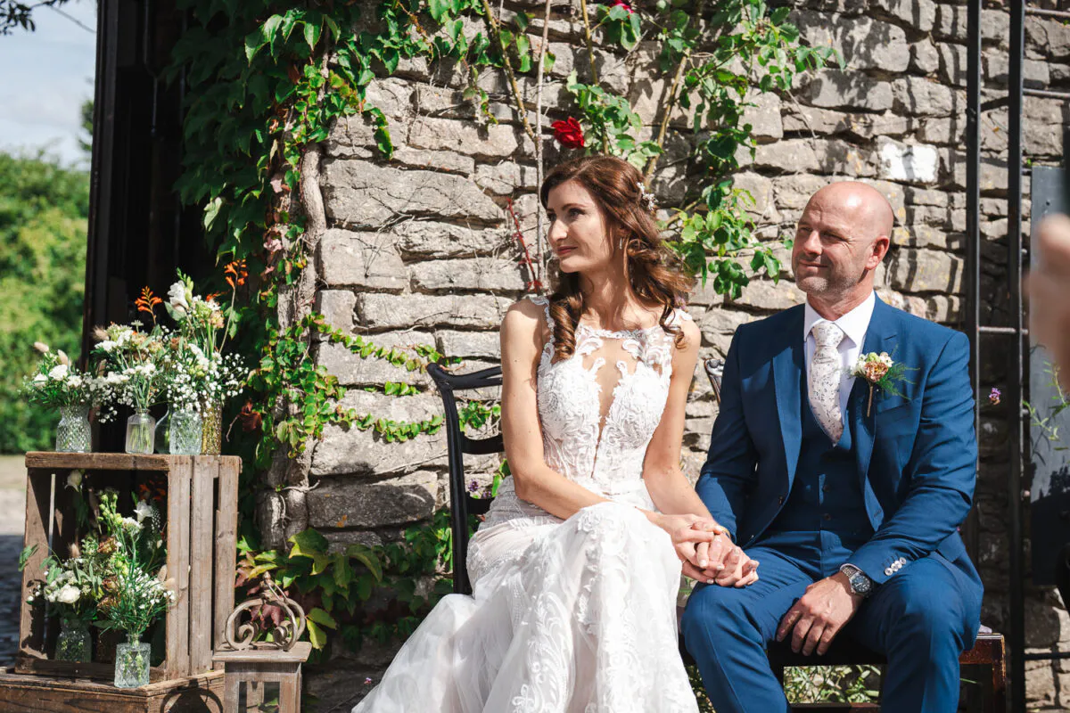 A bride in a detailed lace gown with an illusion neckline and a groom in a sharp blue three piece suit sit together holding hands against a rustic stone wall backdrop.