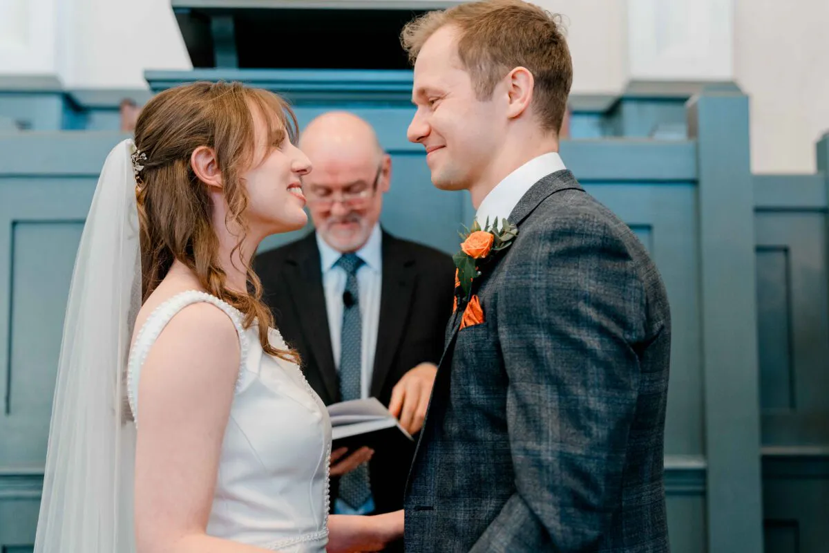 A bride and groom exchange loving glances during their indoor wedding ceremony while a celebrant stands behind them in a blue panelled room.