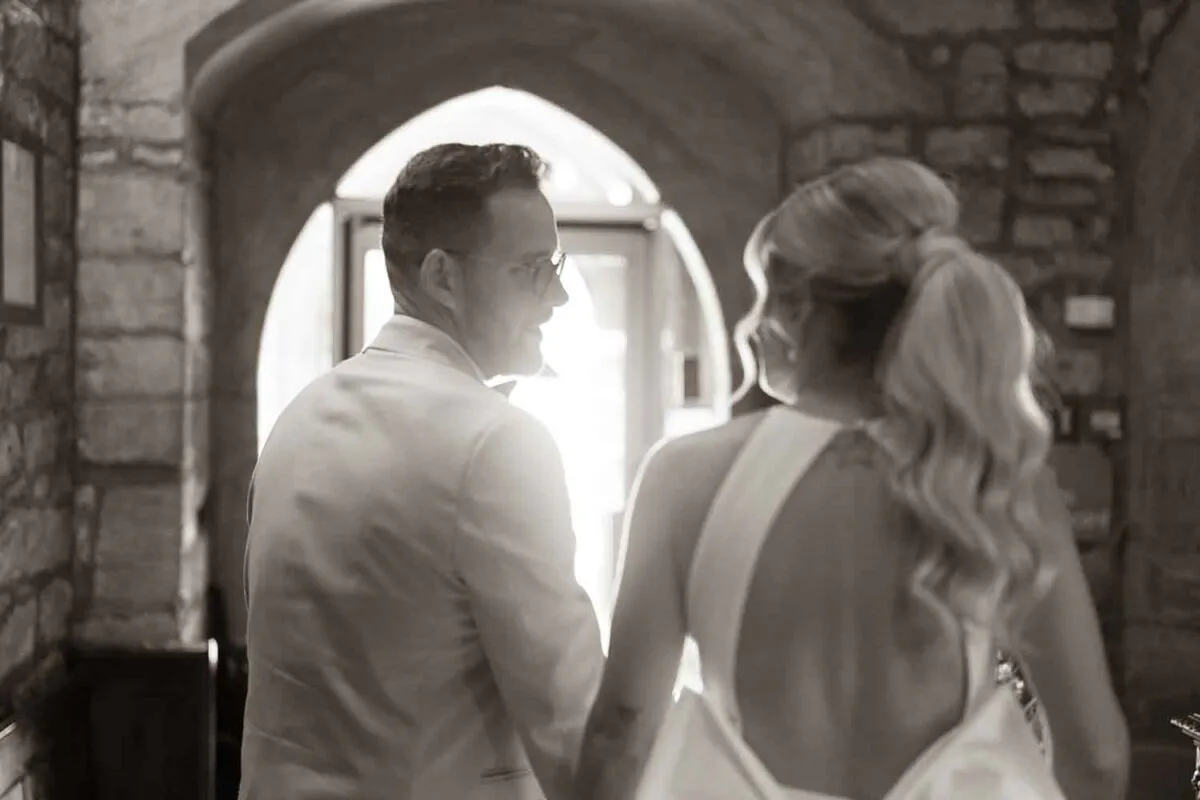 A newlywed couple shares a stolen glance in an over-the-shoulder shot framed by a stone archway, captured in a warm sepia tone.