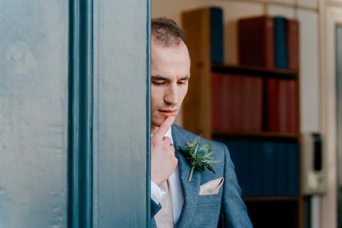 A groom in a grey checked suit with a thistle boutonniere thoughtfully looks through a doorway inside a library.