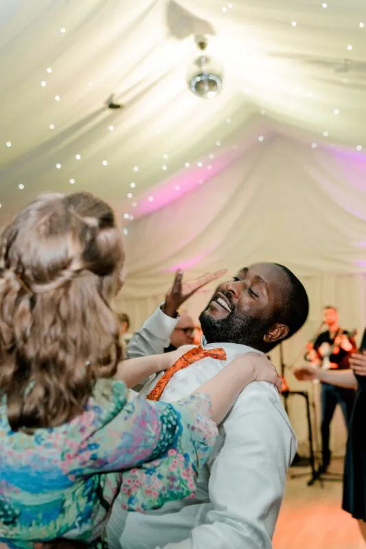 A joyous newlywed couple share a fun moment while dancing under a draped marquee ceiling illuminated by fairy lights and a disco ball.