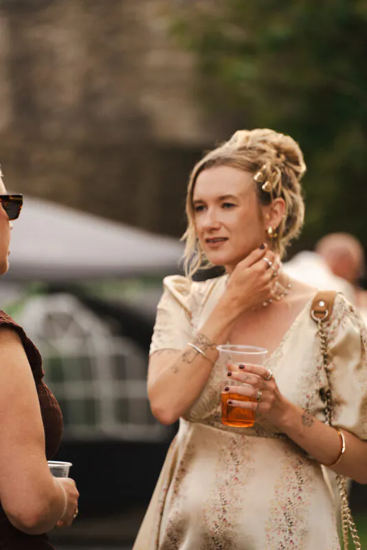 A guest at a wedding drinks reception wearing a floral print dress with puffed sleeves and hair styled in a textured bun holds a plastic cup while chatting outside.