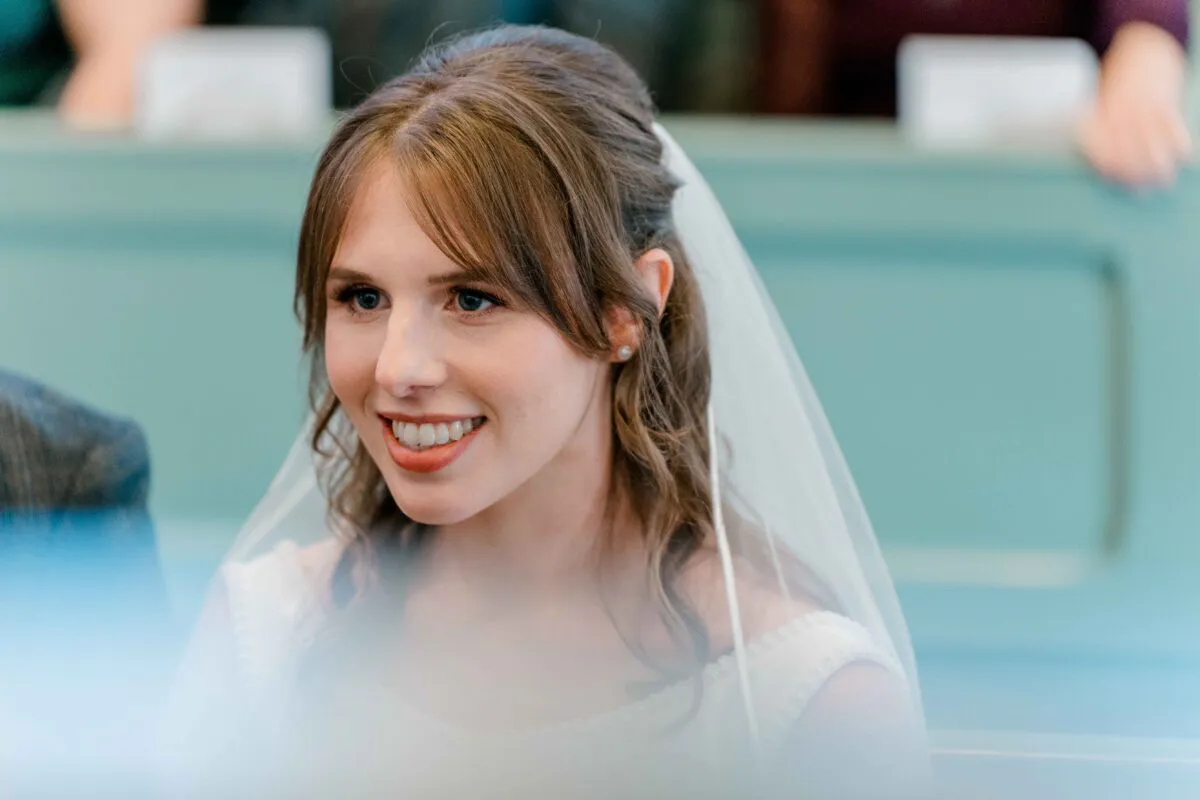 A smiling bride with half-up hair and a veil looks on during a church wedding ceremony.