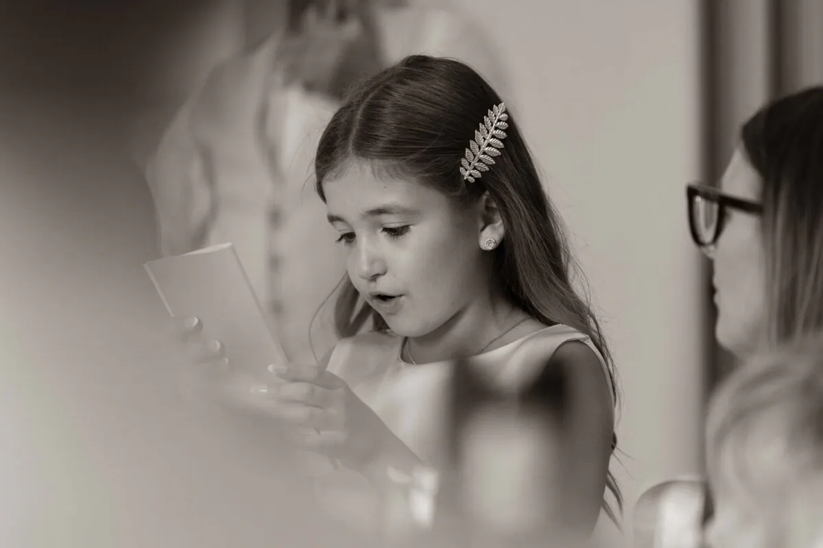 A young bridesmaid with a leafy hair comb attentively reads a ceremony programme during the wedding speeches.