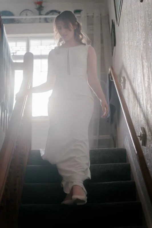 A stunning bride descends a carpeted staircase in a sleek column wedding dress, illuminated by soft natural light streaming through a window behind her.
