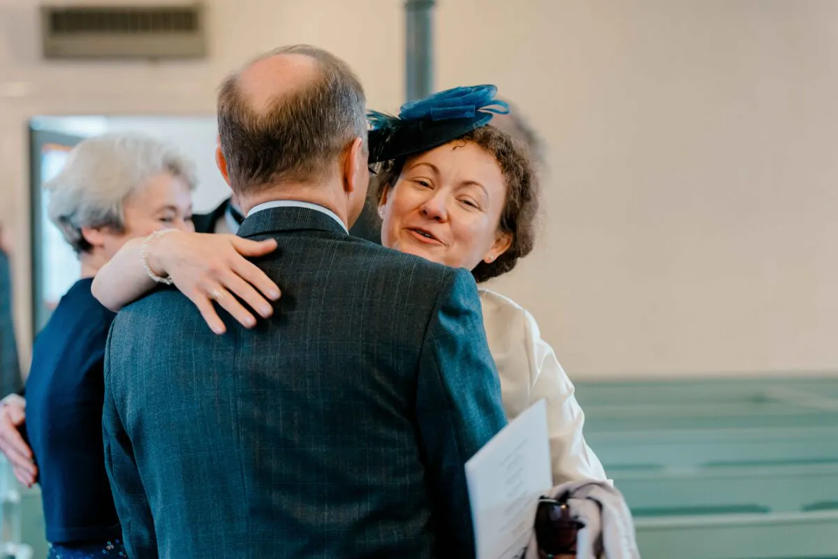 A wedding guest wearing a blue fascinator warmly embraces a man in a checked suit during a reception.