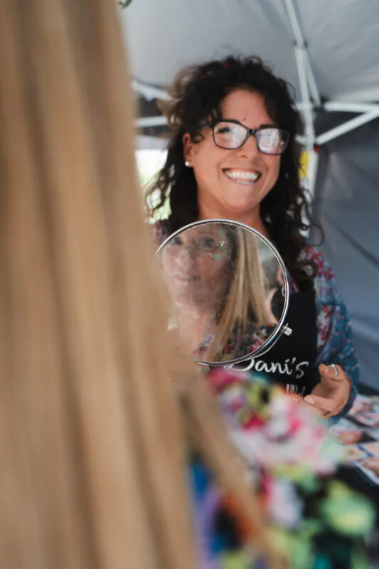 A smiling stylist holds up a round mirror to show a client her makeup reflection during the wedding morning preparations.