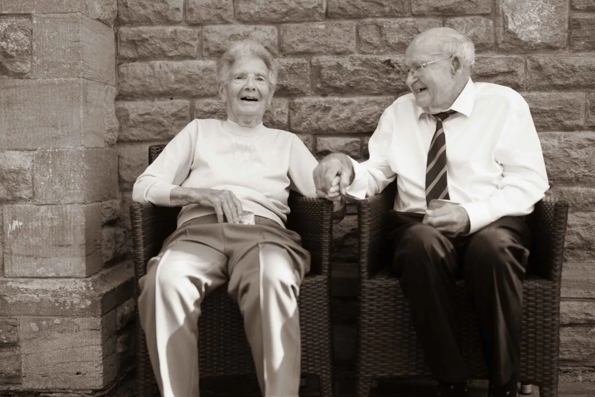 An elderly couple sits together outdoors holding hands in a tender, joyful moment against a rustic stone wall backdrop.