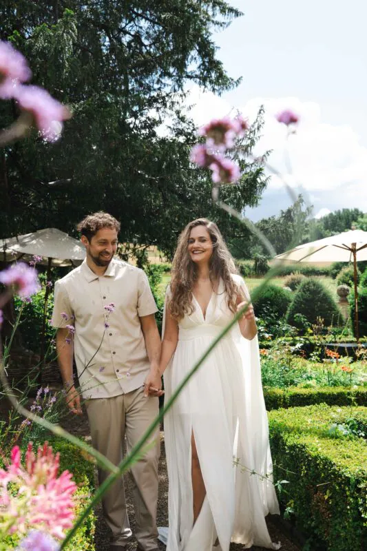 A smiling bride and groom walk hand in hand through an English country garden on a sunny summer day, framed by purple wildflowers.