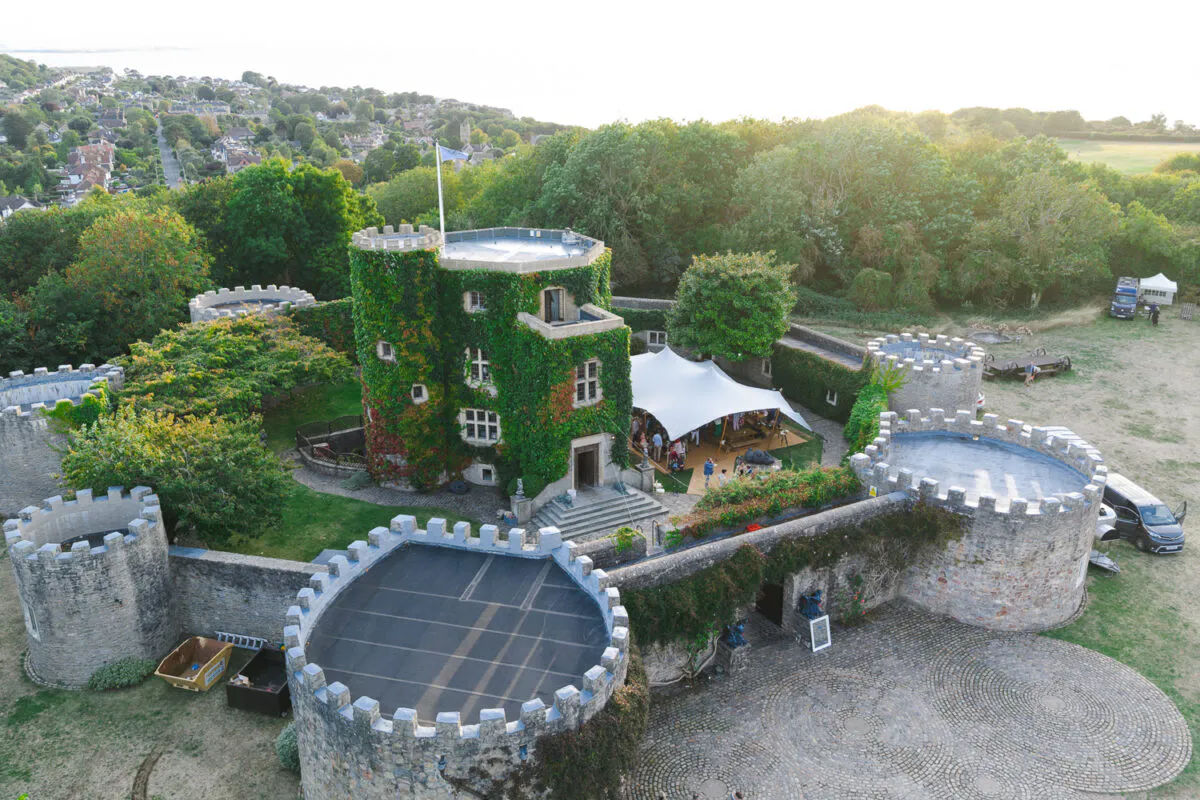 An aerial drone view captures a magnificent castle wedding venue featuring ivy-clad stone towers, a cobbled courtyard, and a white stretch tent hosting guests in the grounds.