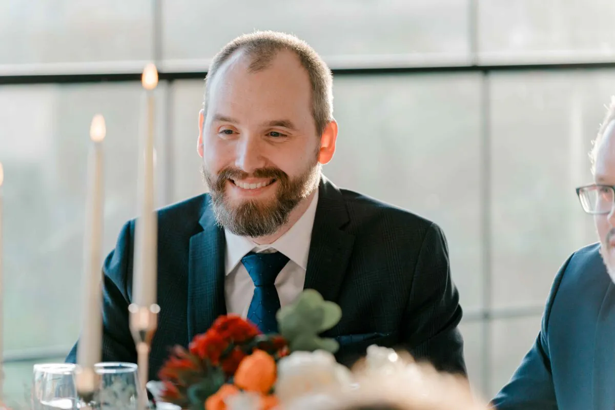A smiling groom sits at the wedding reception table during speeches, framed by lit taper candles and floral centrepieces.