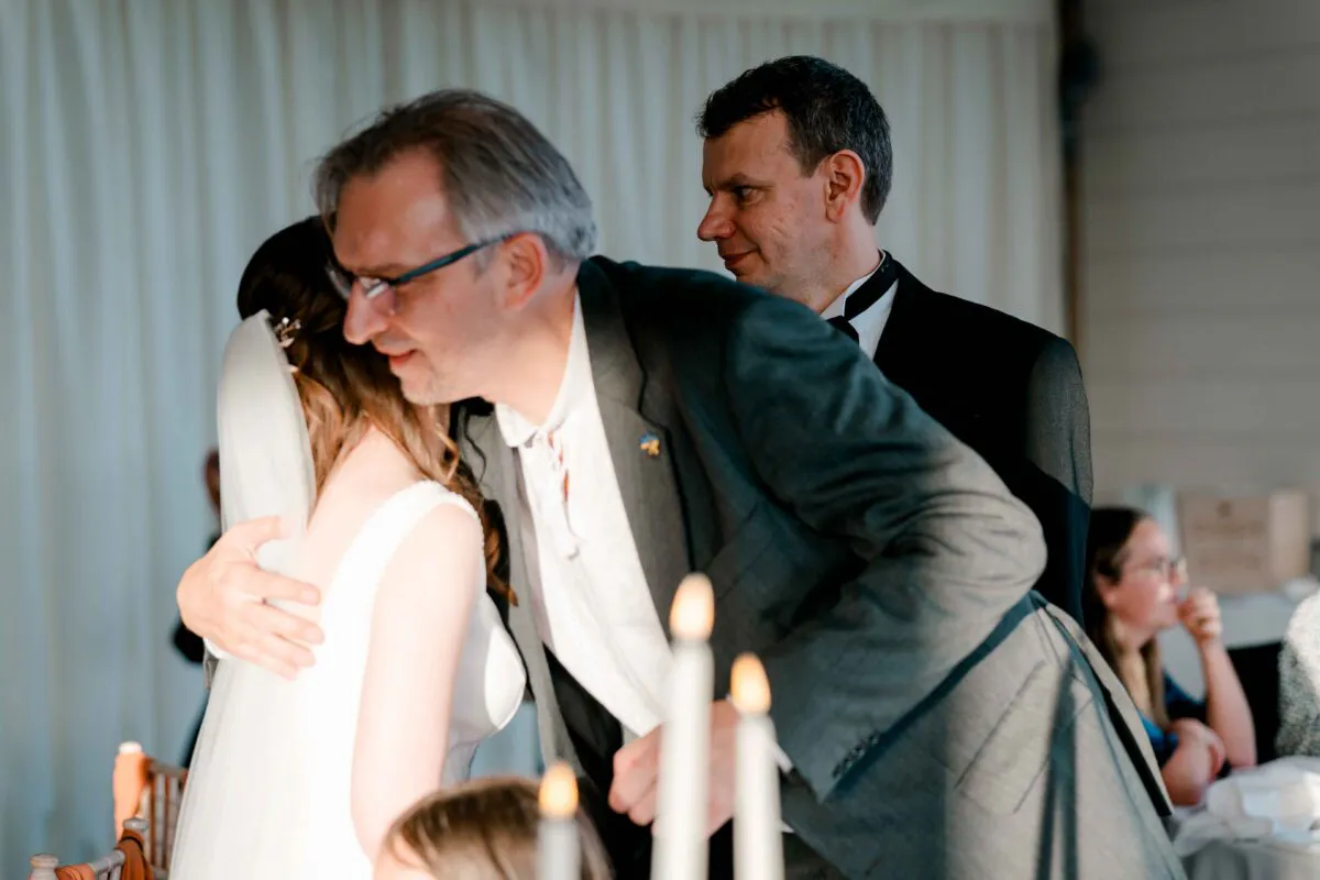 An emotional father of the bride embraces the bride warmly during the wedding reception while guests look on softly in the background.