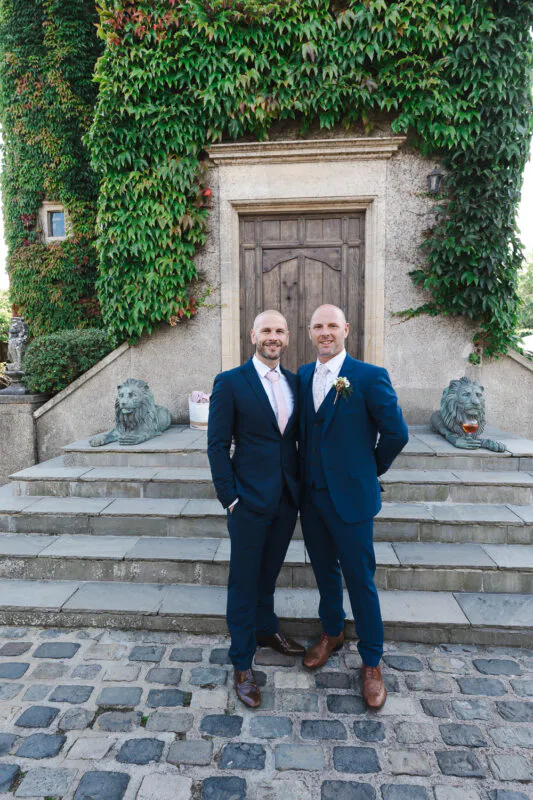 Two grooms in matching navy suits stand smiling in front of an ivy-covered stone building with a wooden door and stone lion statues.
