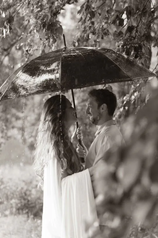 A newlywed couple shares a tender moment smiling at each other beneath a wet umbrella in a wooded area during a gentle rain shower.