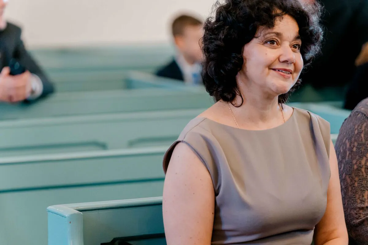 A mature female guest with dark curly hair smiles attentively while seated in a row of pale green pews during a wedding ceremony.