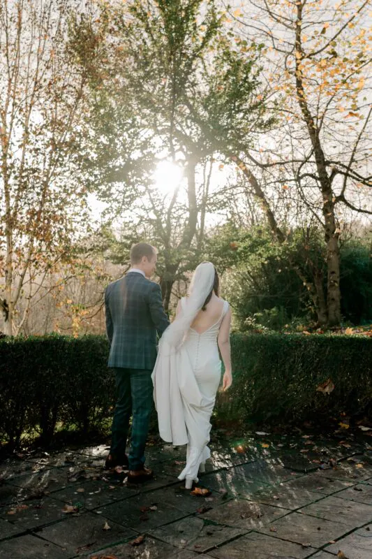 A newlywed couple walks hand in hand along a stone terrace framed by autumn foliage, backlit by the afternoon sun.