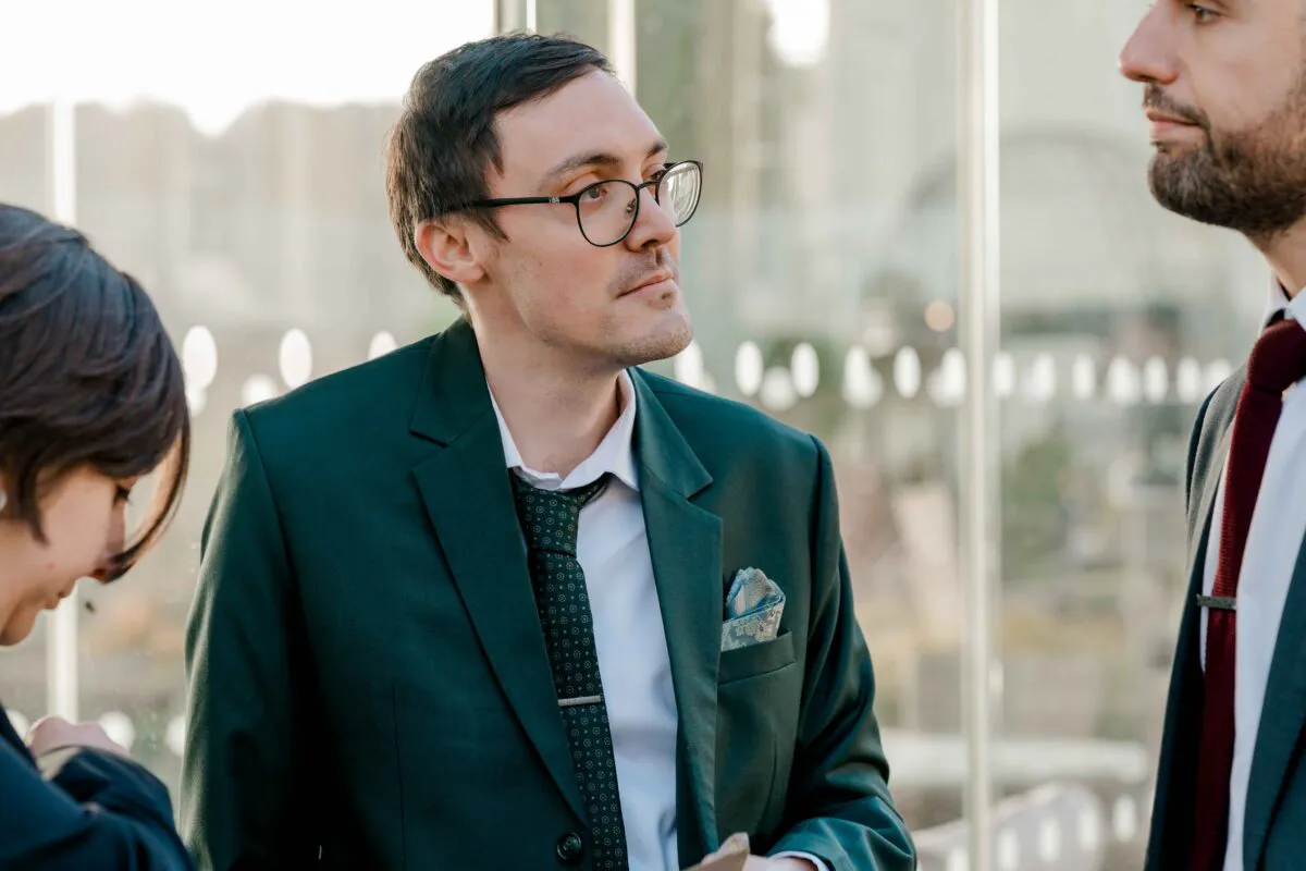A wedding guest in a dark green suit and patterned tie converses with others near a glass partition during a lively reception.