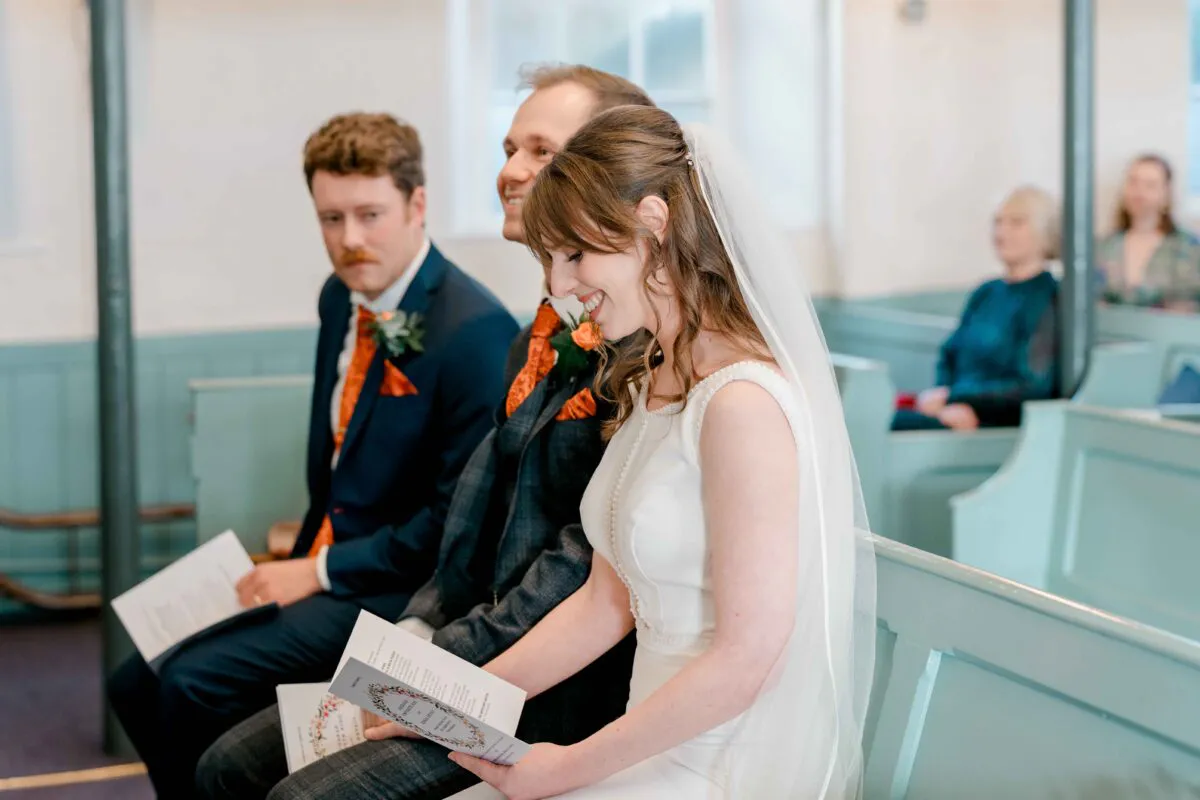 A smiling bride and groom sit together on mint green church pews during a wedding ceremony, holding service programmes while guests look on from behind.
