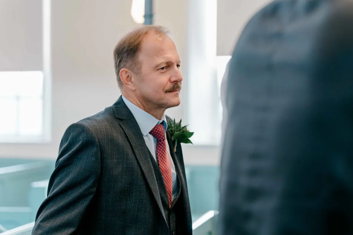 The father of the bride wearing a grey checked suit and patterned red tie looks attentively during the wedding ceremony.