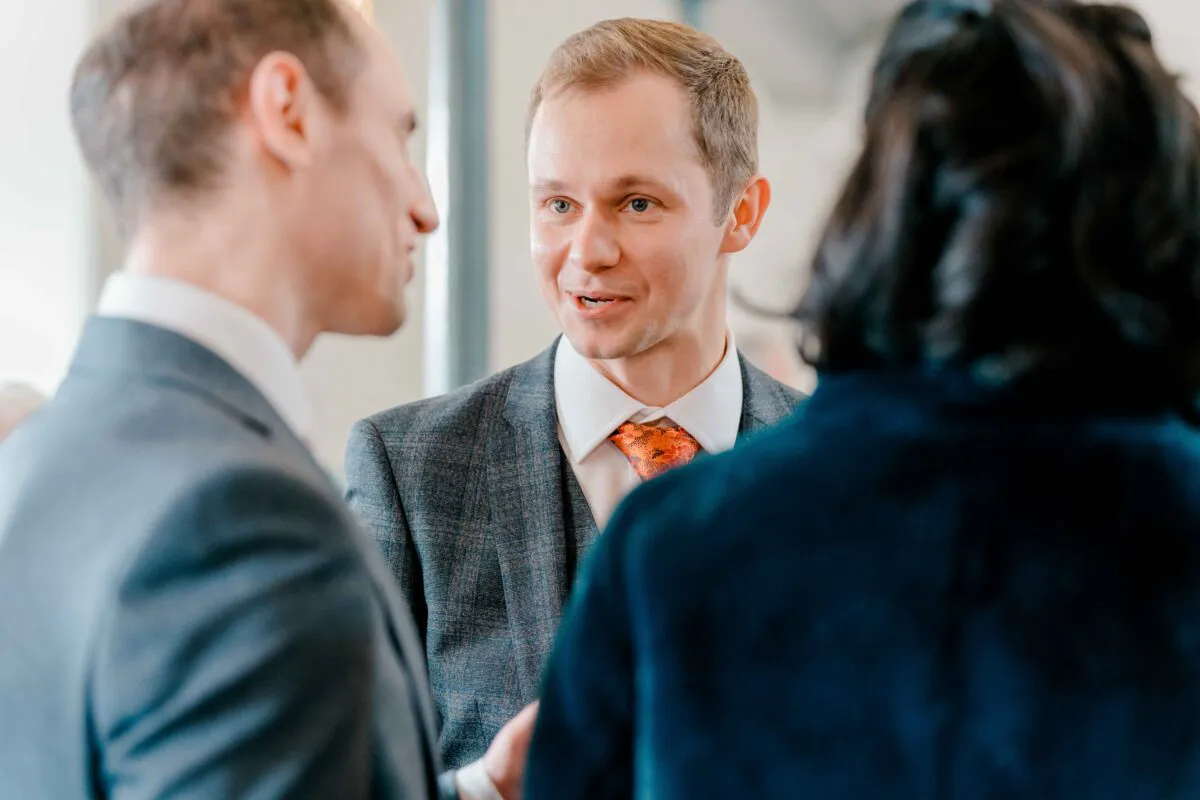 A guest in a grey checked suit and patterned tie converses with other attendees during the wedding reception.