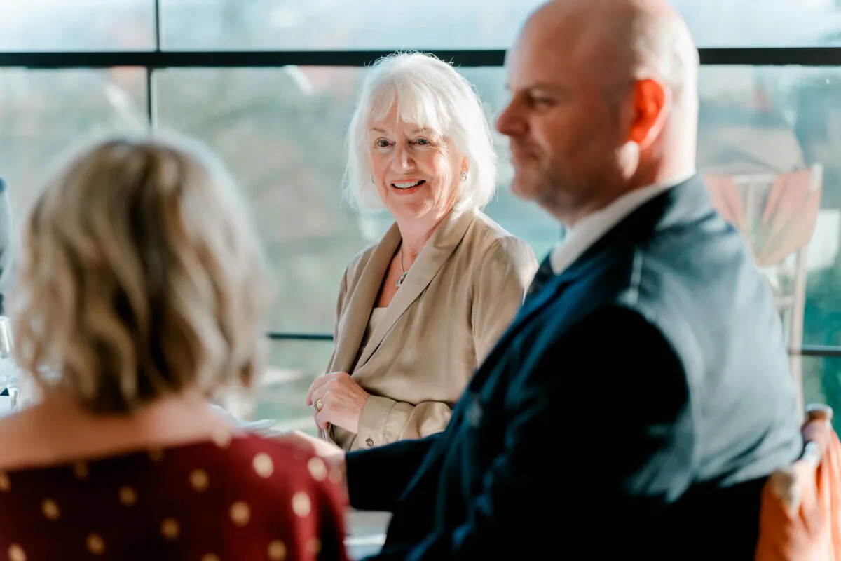 An older woman smiling warmly at a wedding reception while sitting near a large window, with other guests blurred in the foreground.