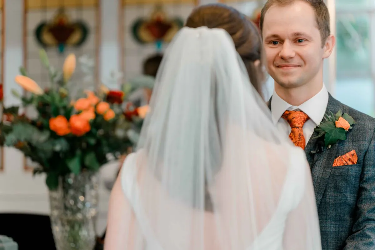 A smiling groom looks lovingly at his bride wearing a veil during their wedding ceremony, with colourful floral arrangements in the background.