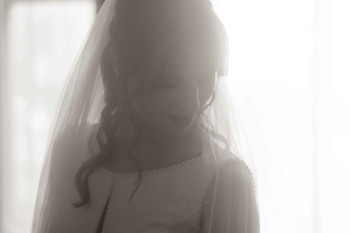 A soft monochrome portrait of a bride looking down thoughtfully while getting ready by a bright window, wearing a delicate veil.