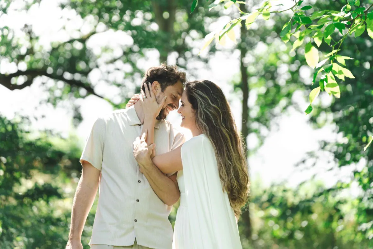 A happy couple share a tender, smiling moment embraced amongst a lush green woodland with dappled sunlight filtering through the trees.