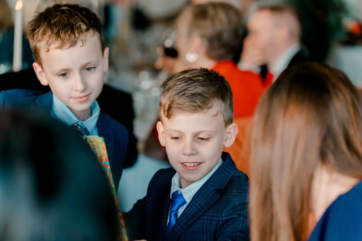 Two young boys in smart checked suits and blue ties look down with amused expressions at a wedding reception.