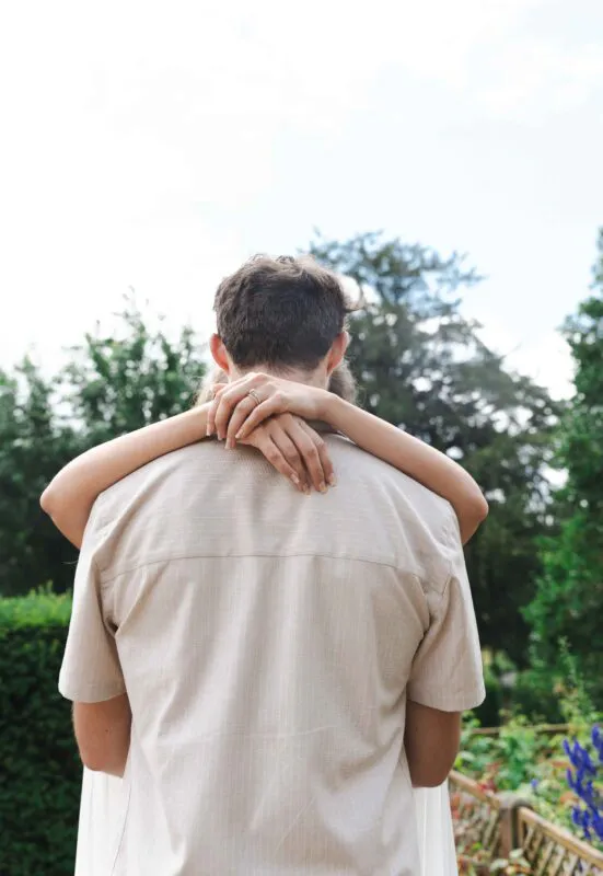 A close-up back view of a couple embracing outdoors, with the partner's arms wrapped around the groom's neck showcasing an engagement ring.