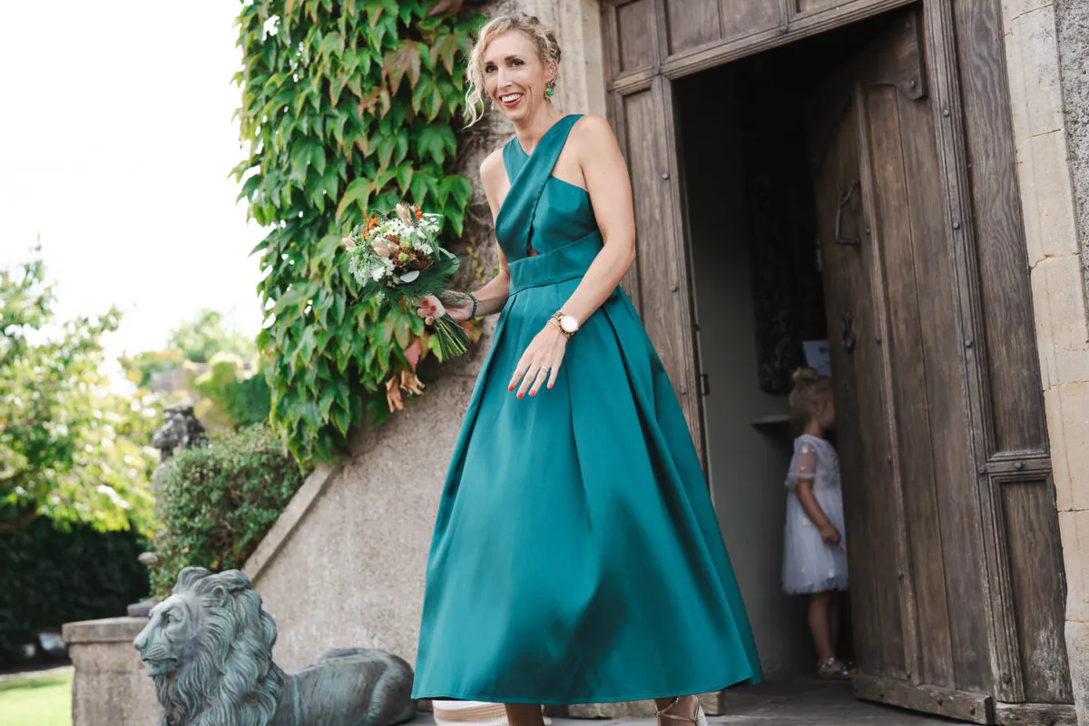 A smiling bridesmaid wearing an emerald green halterneck satin dress and holding a bouquet exits through a wooden doorway next to an ivy-covered stone wall.