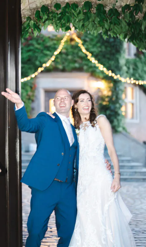 A joyful newlywed couple laugh and wave as they step out through a doorway into a cobbled courtyard illuminated by festoon lights.