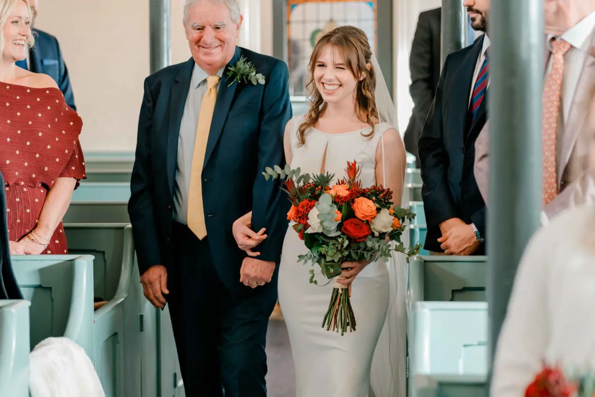 A smiling bride walks down the aisle arm in arm with her father inside a church, holding a vibrant bouquet of orange and red flowers.