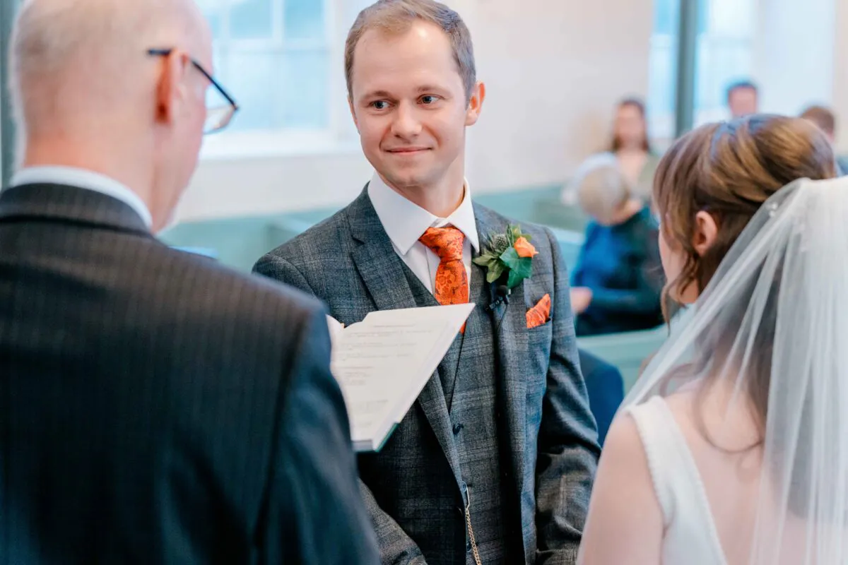 The groom smiles warmly at his bride while standing at the altar during their church wedding ceremony.
