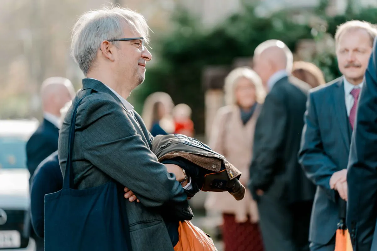 A mature male guest wearing a grey suit and glasses stands with arms crossed while mingling with other wedding attendees outdoors.