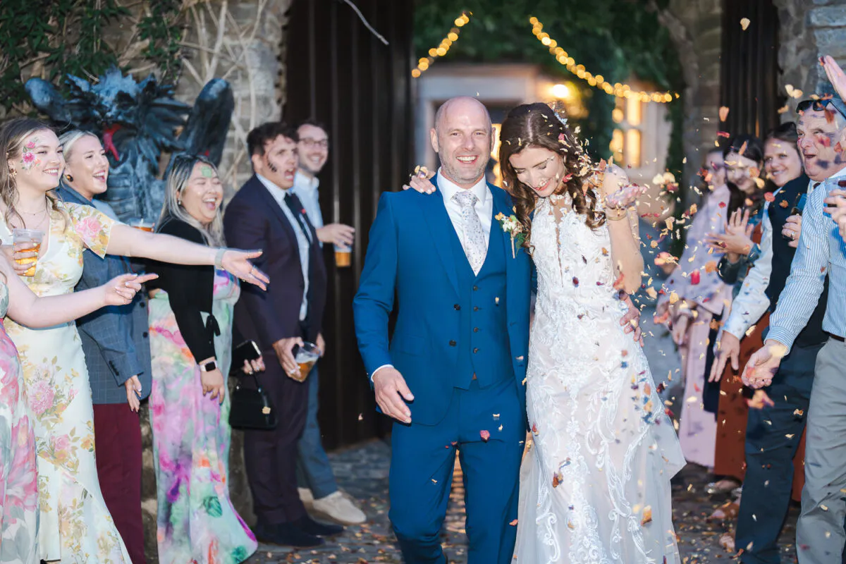 A joyful bride and groom walk hand in hand through an outdoor stone archway while wedding guests joyfully shower them with colourful confetti.