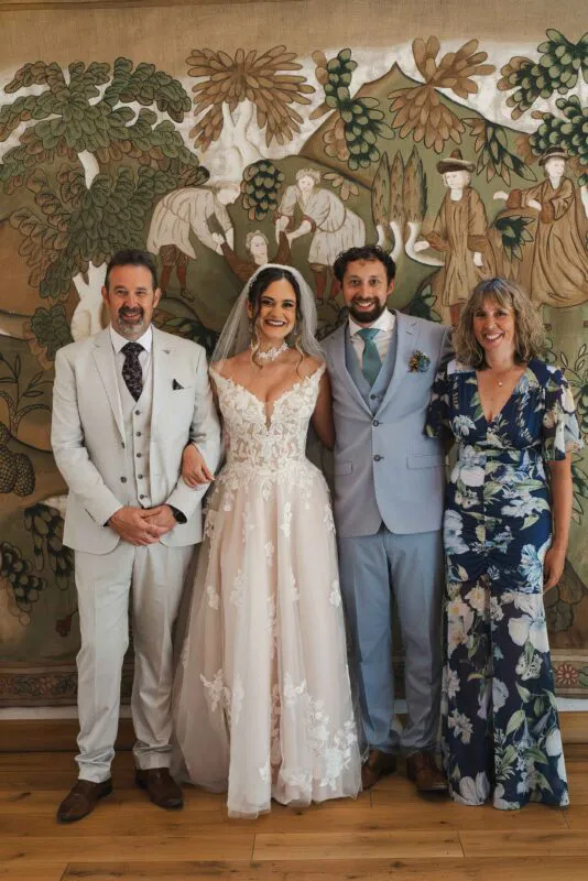 A newlywed couple poses happily alongside family members in front of a large decorative tapestry inside a venue room.