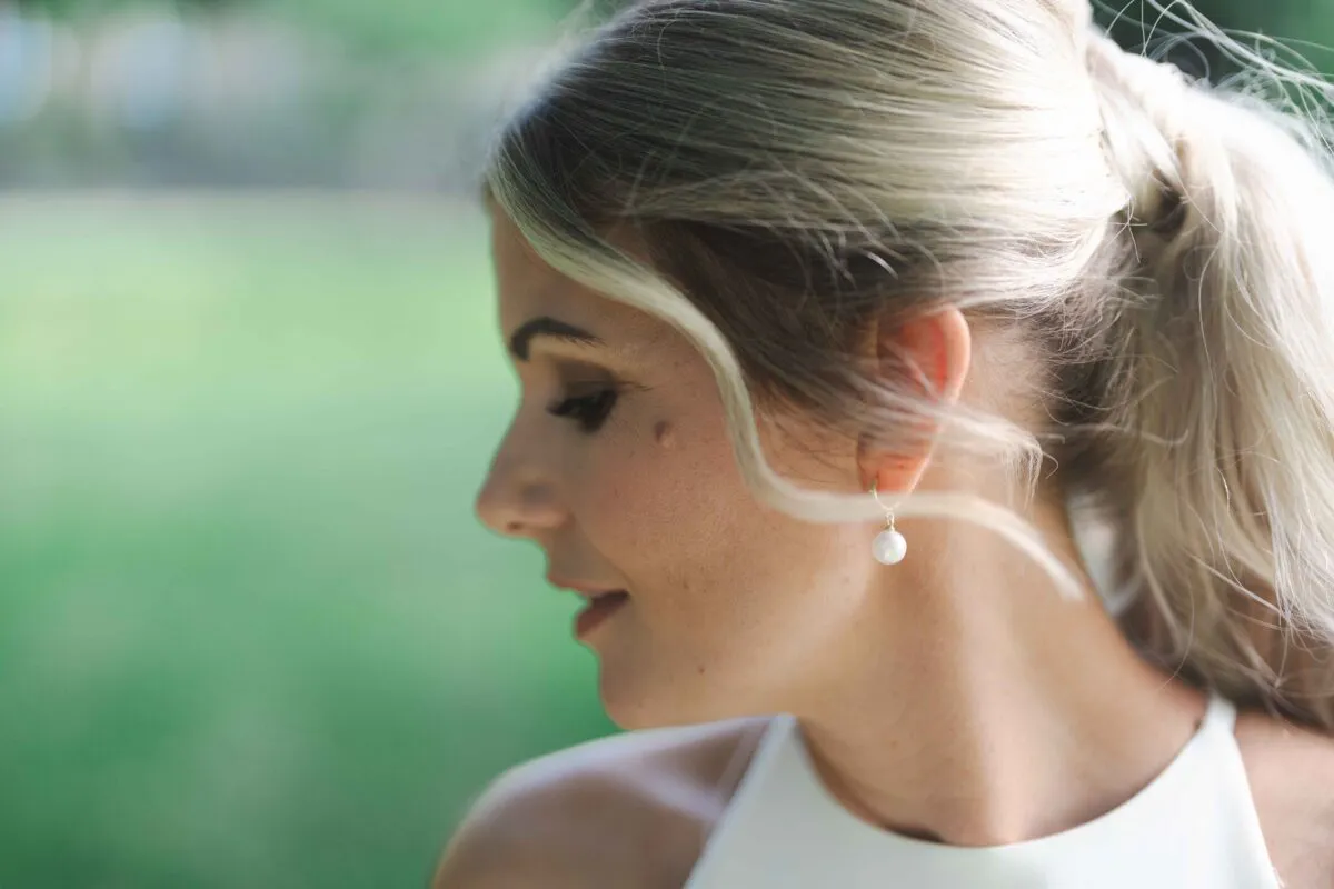 A close-up profile portrait of a blonde bride wearing delicate pearl drop earrings and a sleek high ponytail against a soft green background.