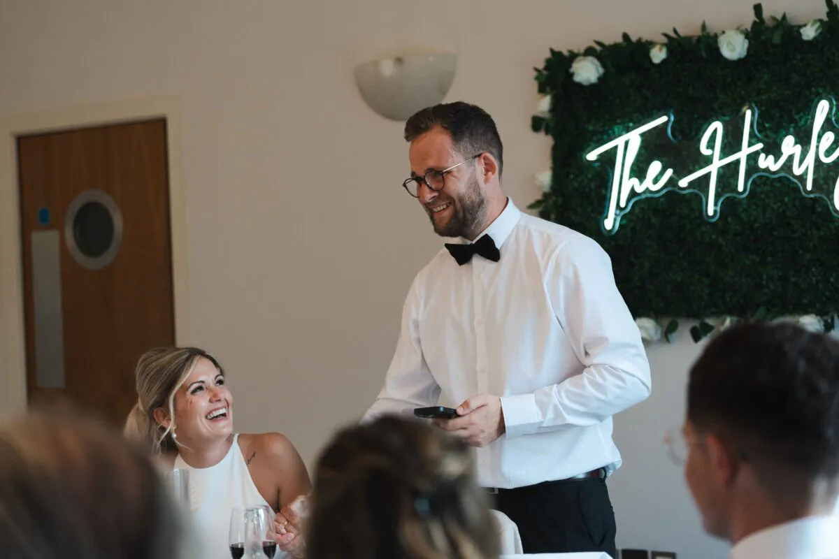 A groom in a white shirt and black bow tie delivers a speech while his laughing partner looks up at him warmly during the wedding reception.