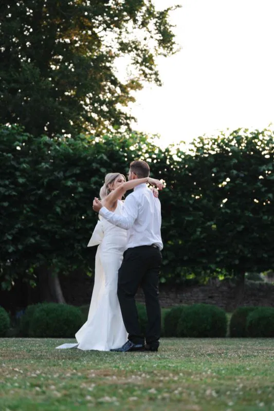A newlywed couple shares an intimate dance outdoors in a manicured garden during golden hour.