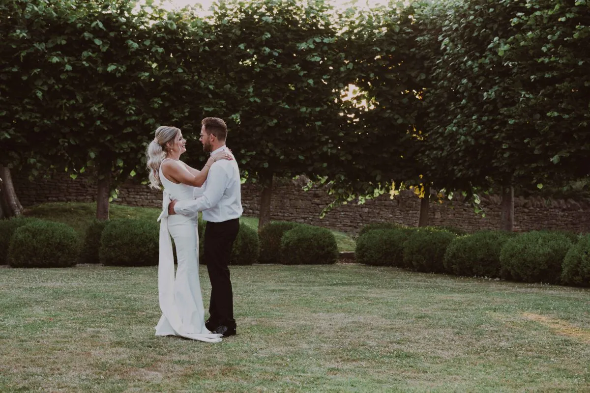 Newlyweds lovingly embrace and smile at each other during golden hour in a scenic garden lined with manicured topiary and mature trees.