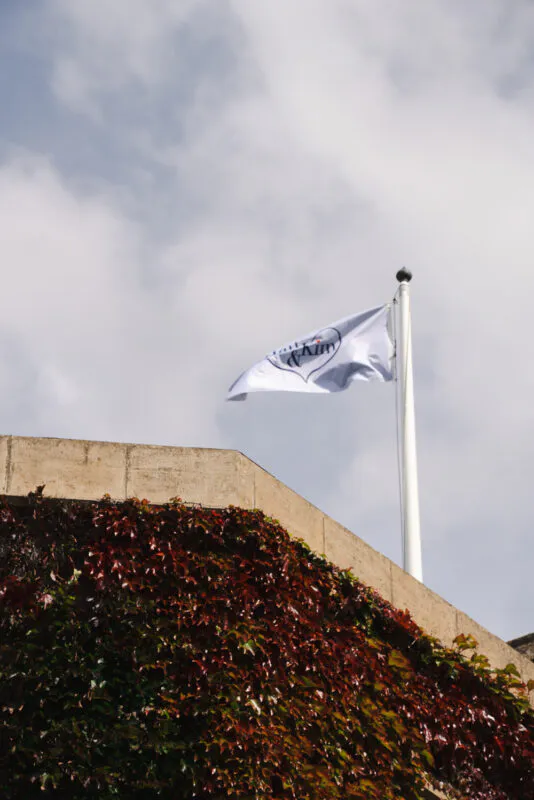 A custom wedding flag flies on a flagpole against an overcast sky, positioned above a stone wall covered in deep red autumn ivy.