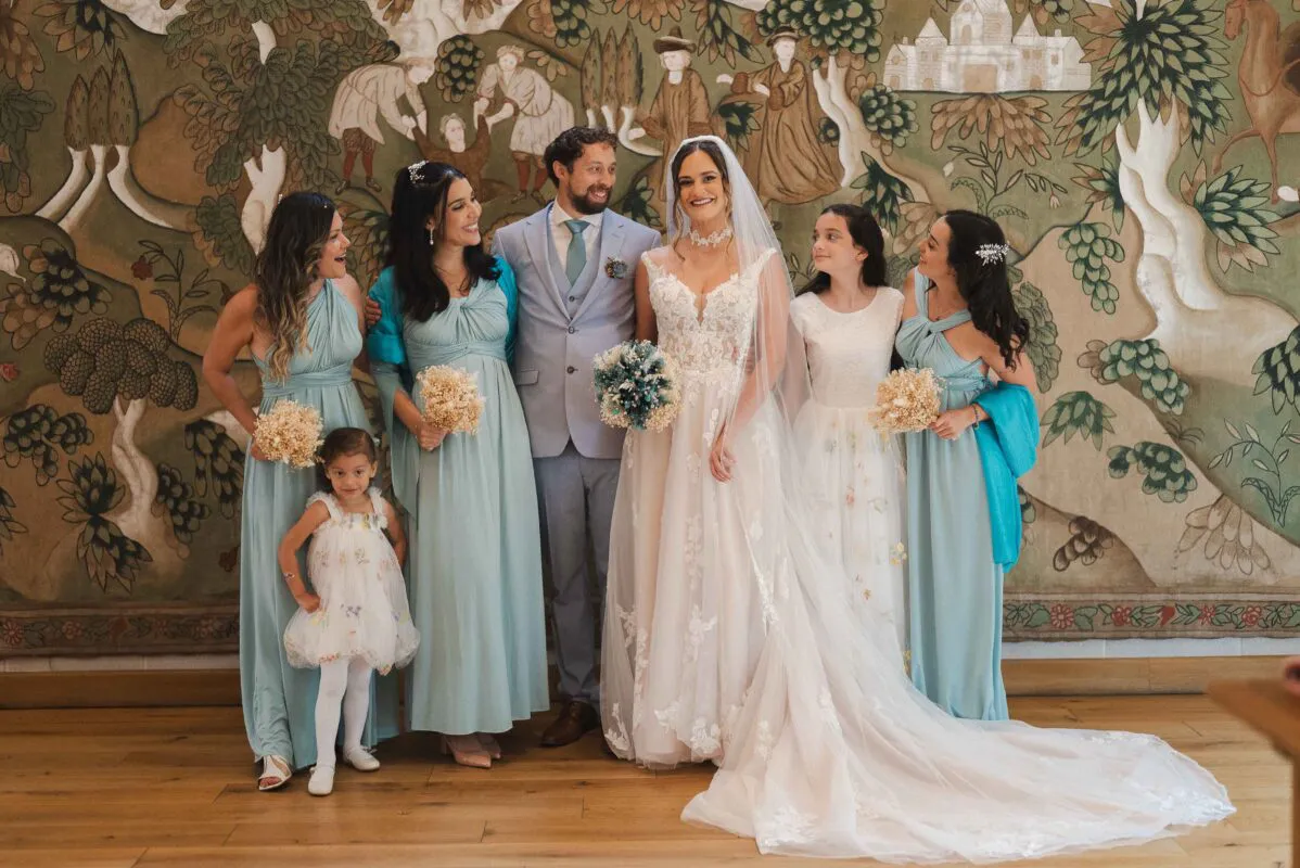 The bride in an appliqued a-line gown and long veil poses alongside her groom in a light blue suit, flanked by bridesmaids in sage gowns and young flower girls in front of an ornate historical tapestry.