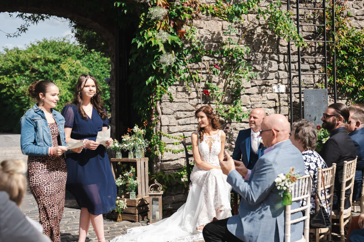 A bride in a detailed lace gown sits seated outside a stone barn during a wedding ceremony while guests watch and laugh.