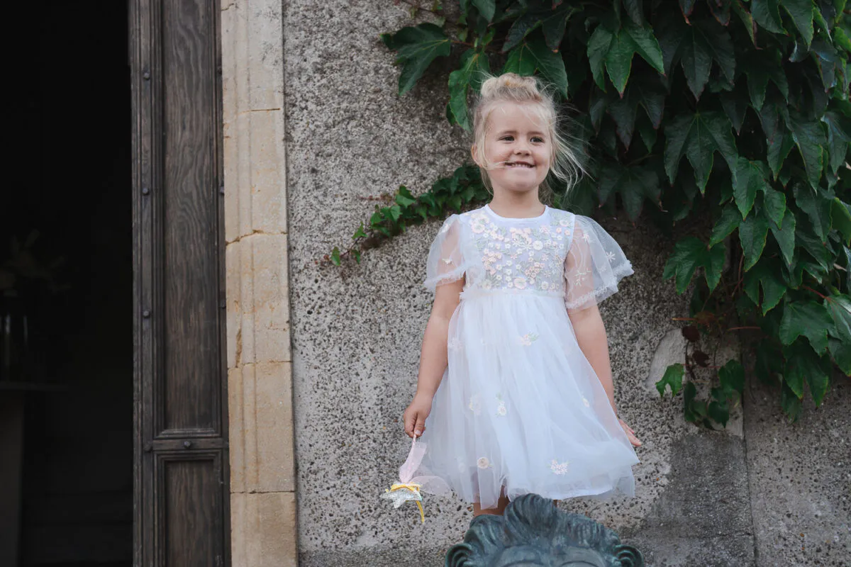 A smiling flower girl wearing a tulle dress with floral embroidery stands happily against a stone wall covered in ivy outside a venue.