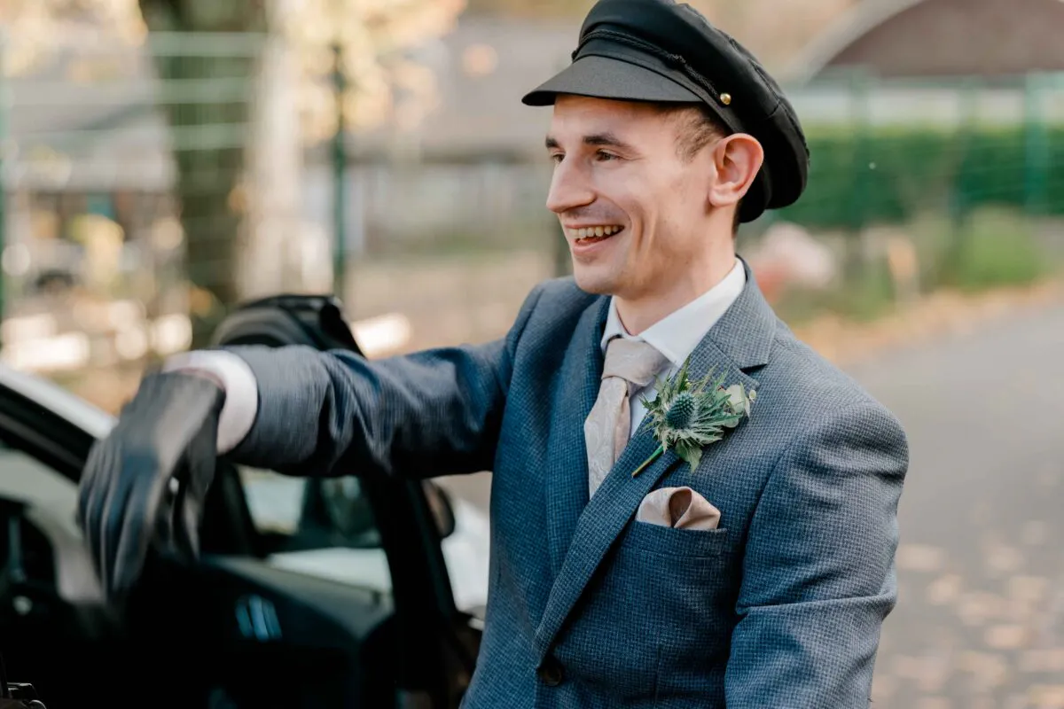 A smiling groom wearing a grey suit, patterned tie, thistle buttonhole, and a chauffeur hat stands next to a wedding car.