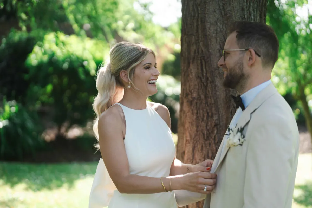 A newlywed couple laughing together outdoors in a sunlit garden, with the bride wearing a sleek halter-neck gown and high ponytail while adjusting the groom's lapel.