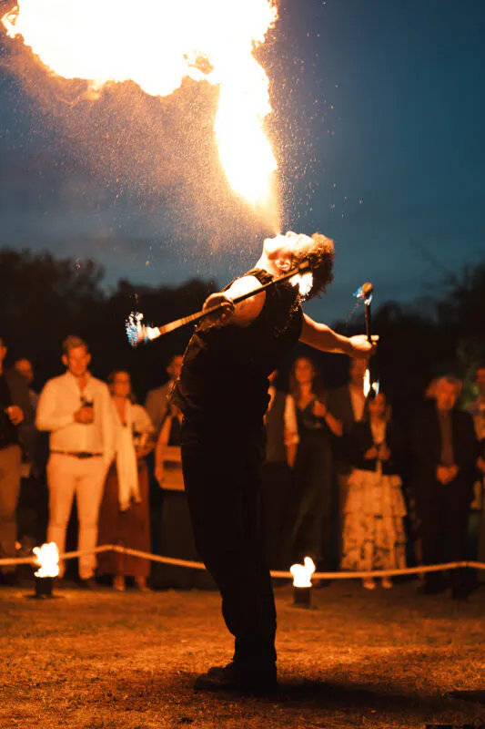 A dramatic nighttime wedding entertainment scene featuring a fire performer breathing a massive plume of fire in front of an astounded crowd of guests.